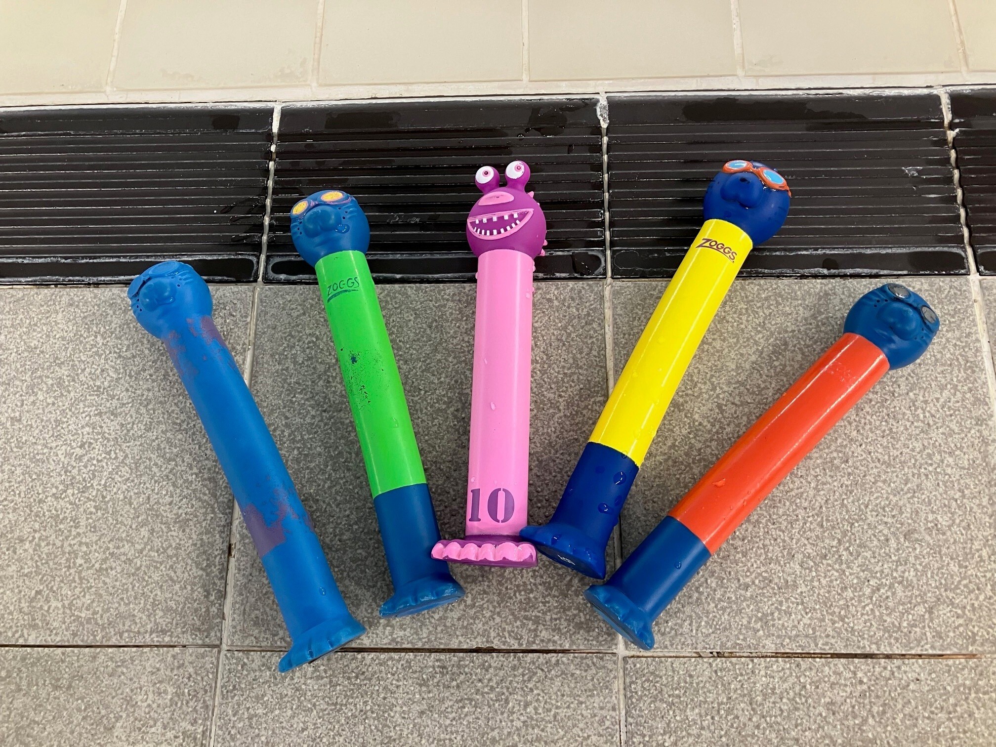 Five colourful pool dive sticks arranged in a fan shape on wet tiles. Each stick has a cartoon character face on top, with colours including blue, green, pink, yellow, and orange. The pink stick features a monster face with two eyes and teeth.