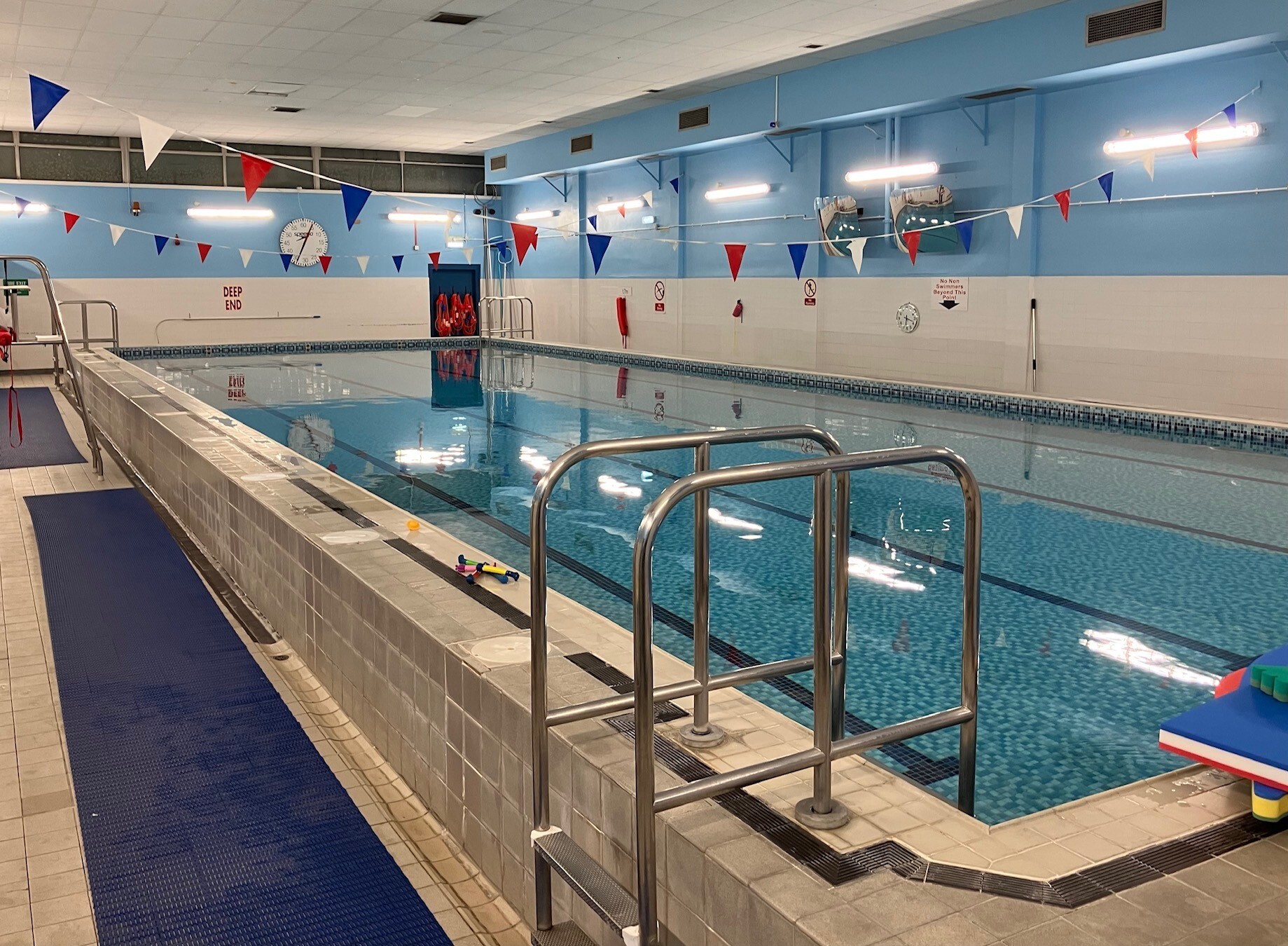 Indoor swimming pool with a tiled surround and steps to enter the pool. The pool features lane markers and there are colourful flags hanging from the ceiling.