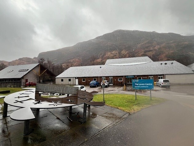 A building with a parking area situated in front, prominently displaying an artistic sculpture in the foreground, with a hill in the background.