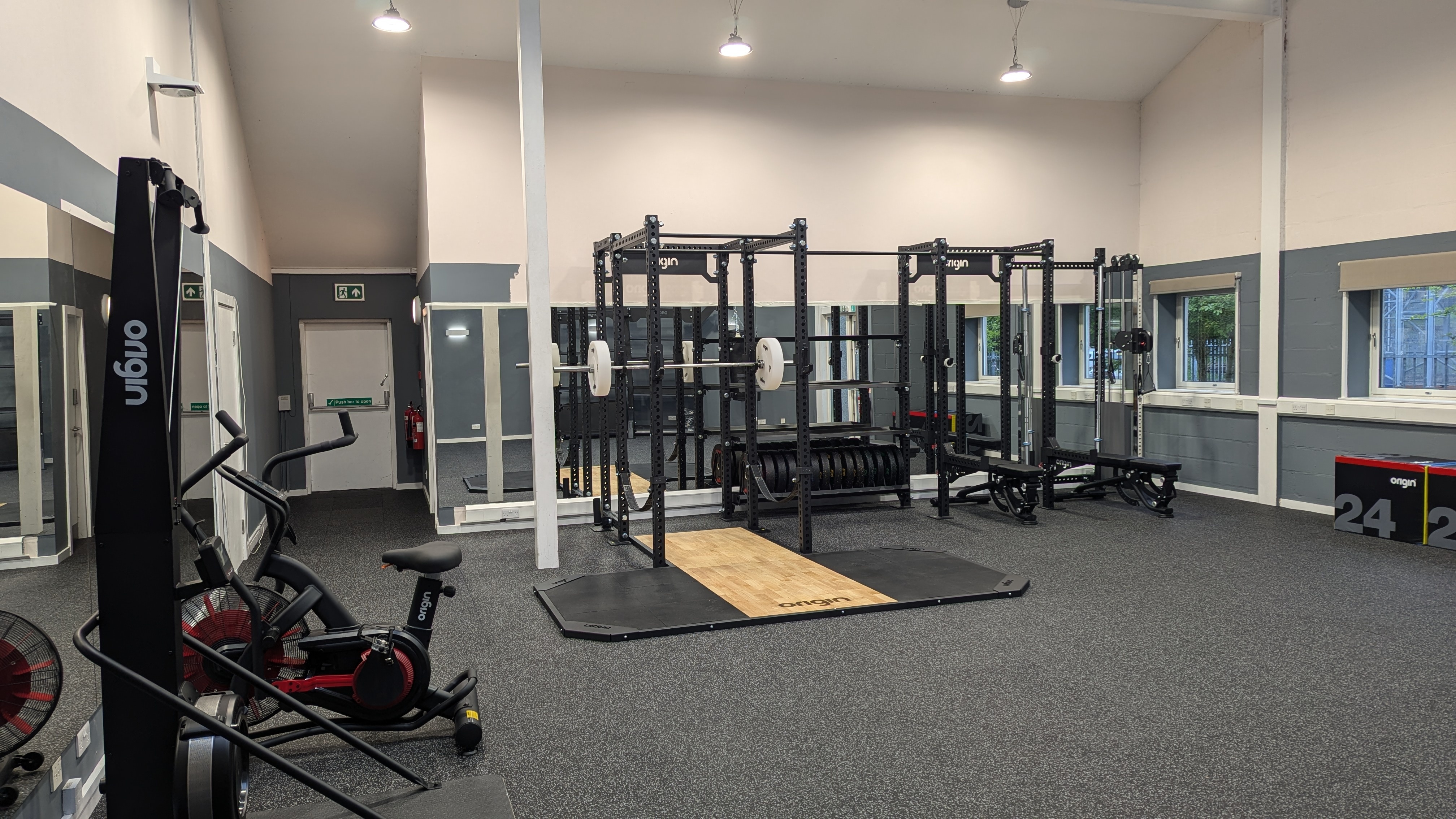 Spacious strength‑training area with black Origin power racks positioned on wooden lifting platforms. A stationary air bike is visible on the left, and plyometric jump boxes are stacked on the right. The room has speckled rubber flooring, grey and cream walls, mirrors along one side and high ceiling lights illuminating the space.