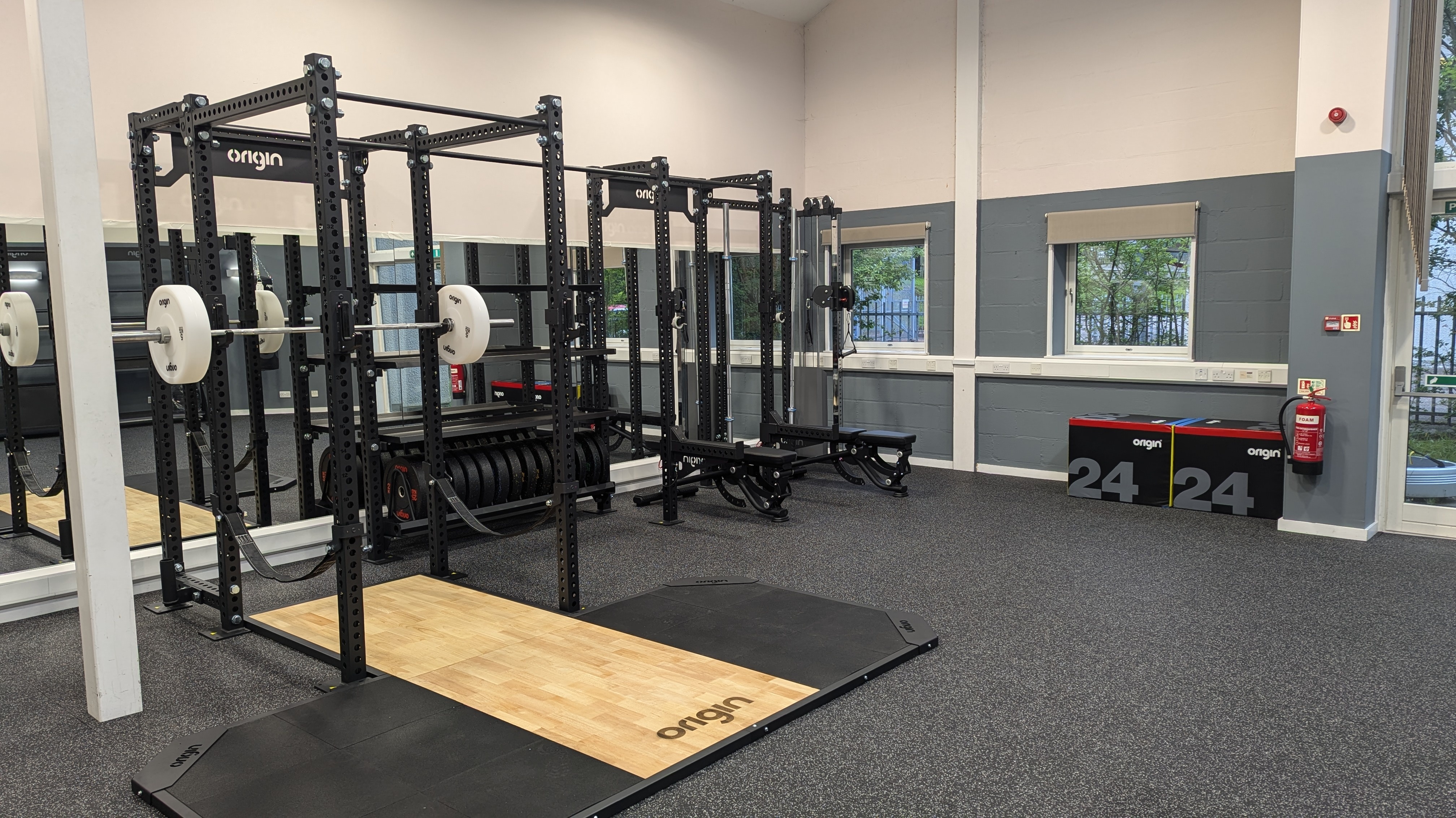 Strength‑training area featuring two black Origin power racks with barbells and white weight plates, positioned on wooden lifting platforms. The room has speckled rubber flooring, grey and cream walls, and windows along the side. Soft plyometric boxes are stacked against the wall, and mirrors line the back wall.