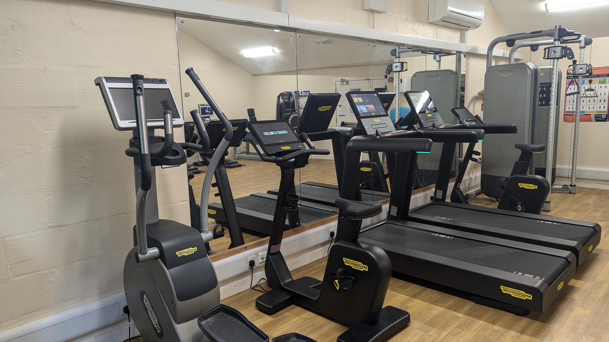 Gym room with light wood flooring featuring Technogym cardio equipment, including an exercise bike, cross-trainer, and two treadmills positioned in front of a large mirrored wall. A strength‑training machine and instructional wall chart are visible in the background, with bright ceiling lights illuminating the space.