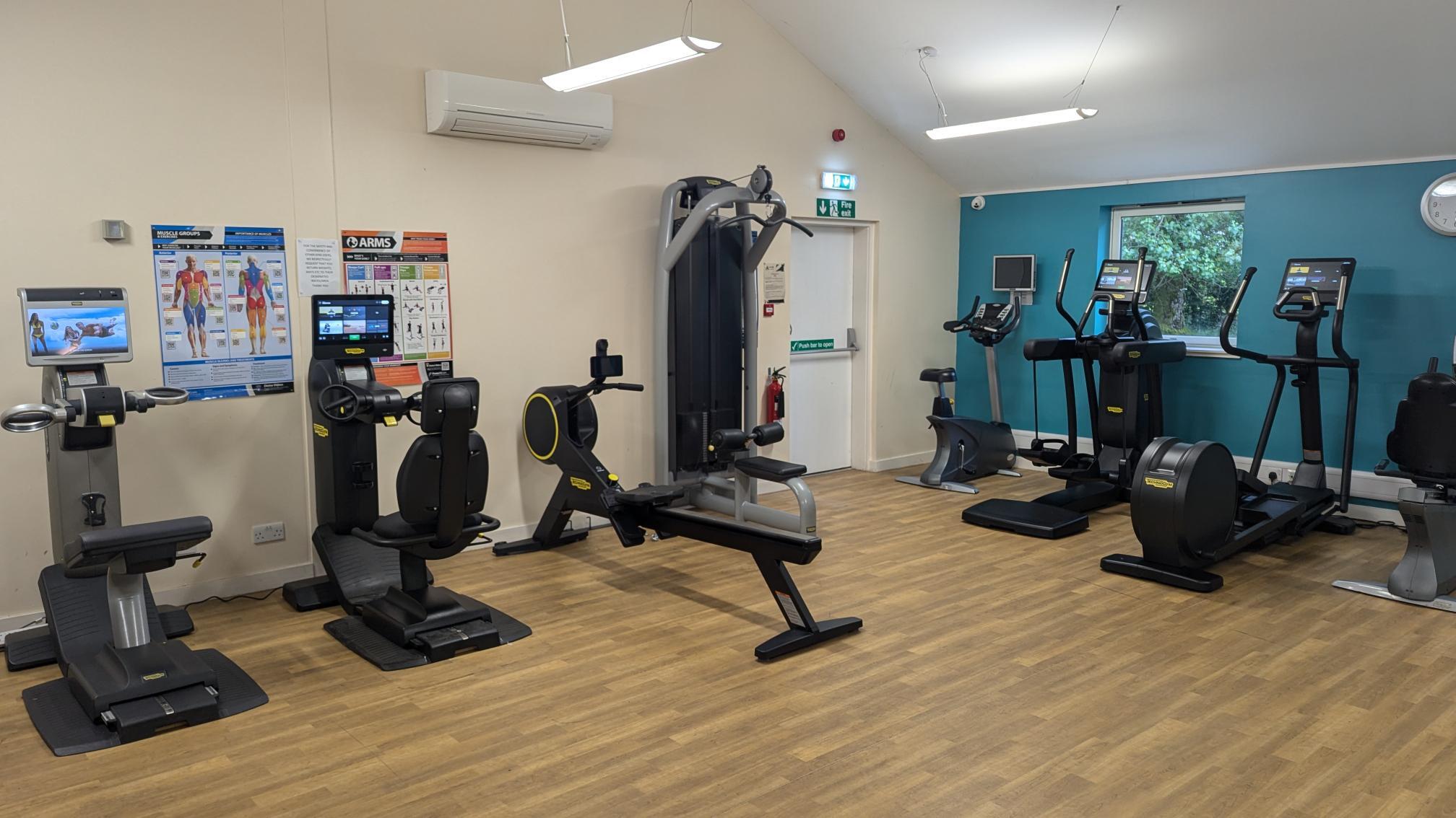 Small gym room with light wood flooring and a variety of Technogym equipment, including a recumbent bike, upright bike, rowing machine, cable station, and two cross-trainers. Posters showing muscle groups are on the wall, and a teal accent wall with a window is on the right. Ceiling lights and natural light brighten the space.