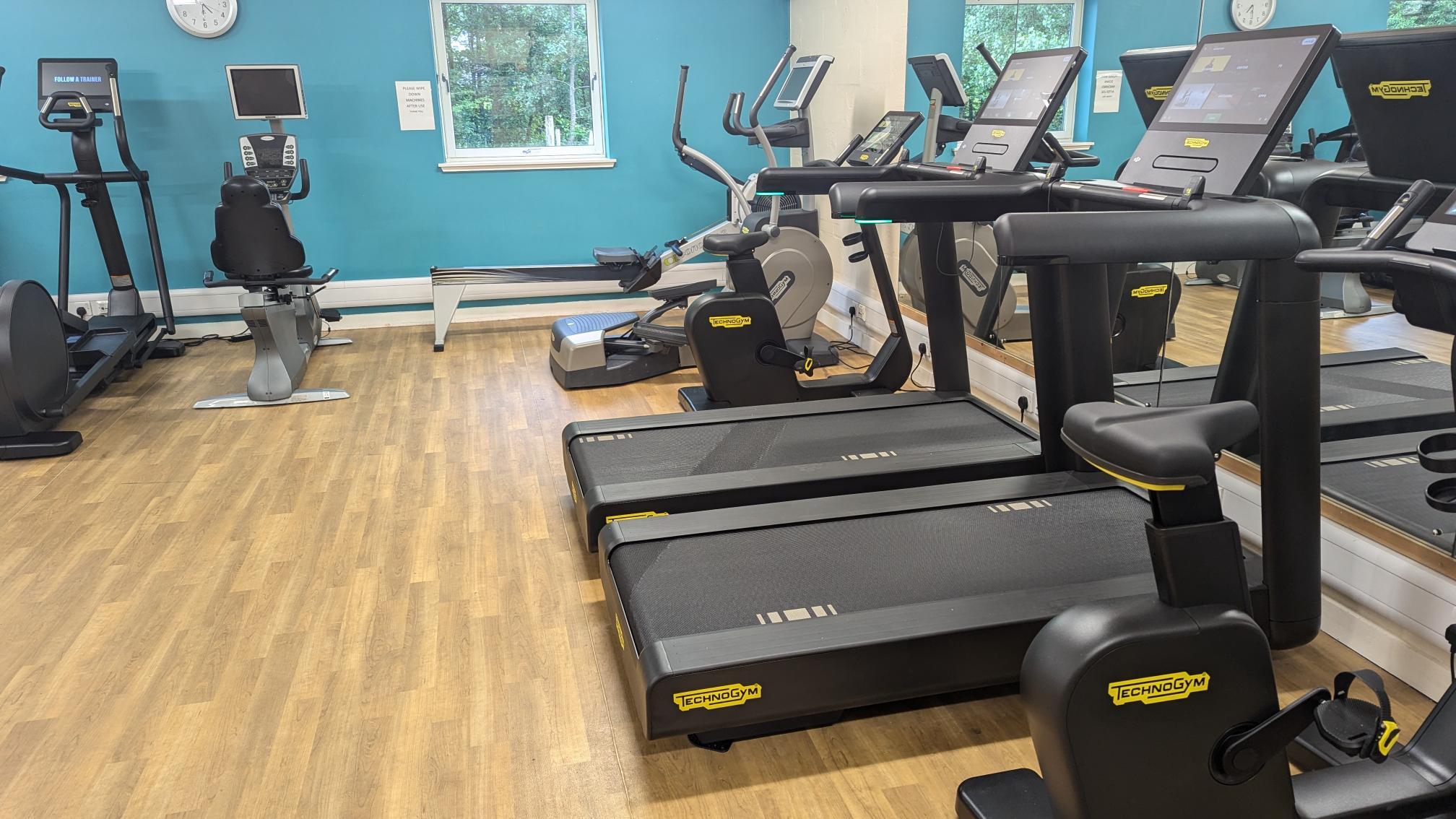 Small gym with light wood flooring featuring Technogym treadmills, cross-trainers, and a recumbent bike arranged along mirrored walls. A teal feature wall with a window is visible in the background, and natural light comes in from outside. The equipment is spaced neatly, creating an open and tidy workout area