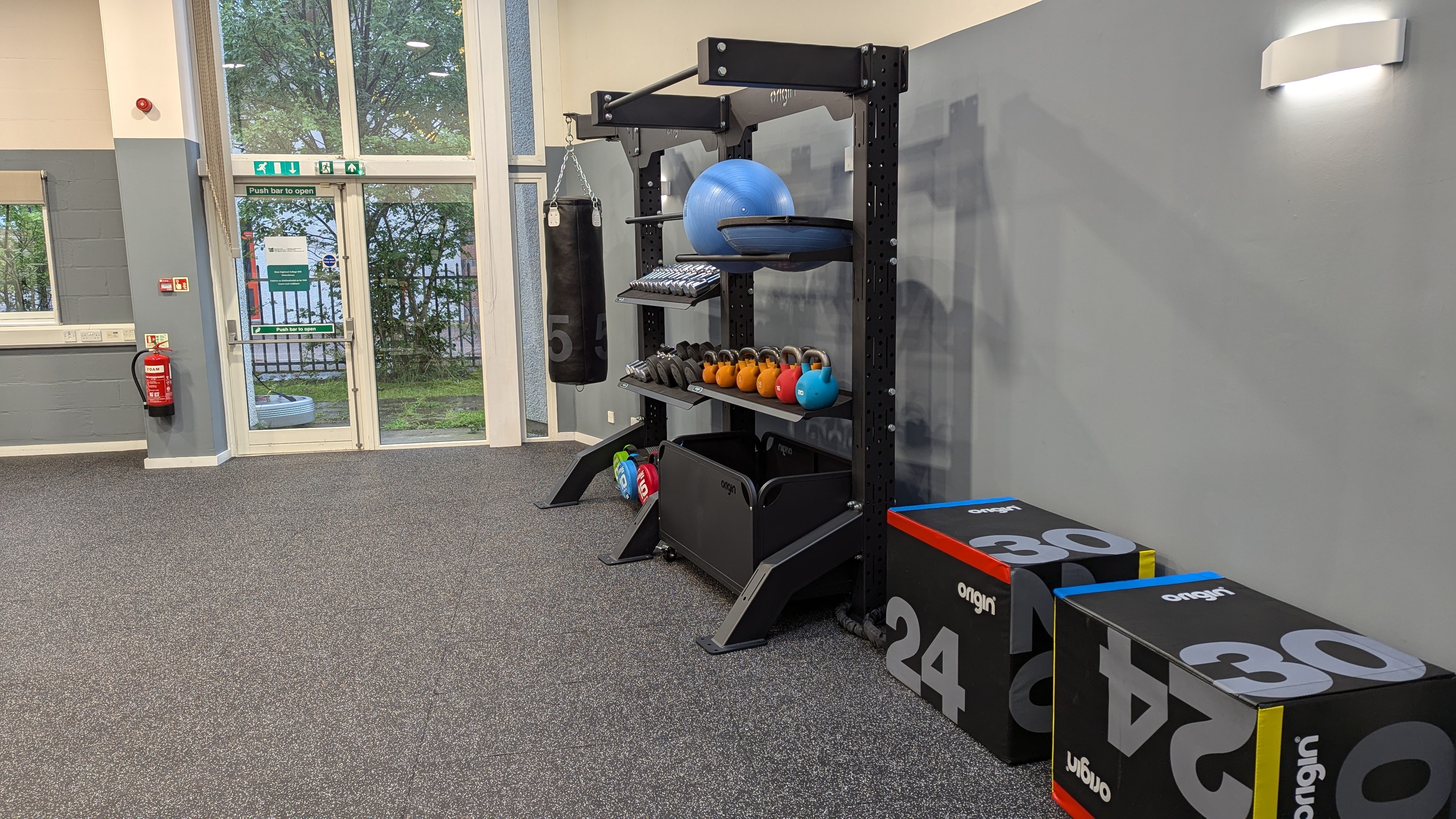 Functional training area with a black storage rack holding kettlebells, medicine balls, and stability balls. A small hanging punch bag is attached to the frame. Soft plyometric jump boxes labeled with different heights are stacked nearby. The room has grey walls, speckled rubber flooring, and large floor‑to‑ceiling windows letting in natural light.