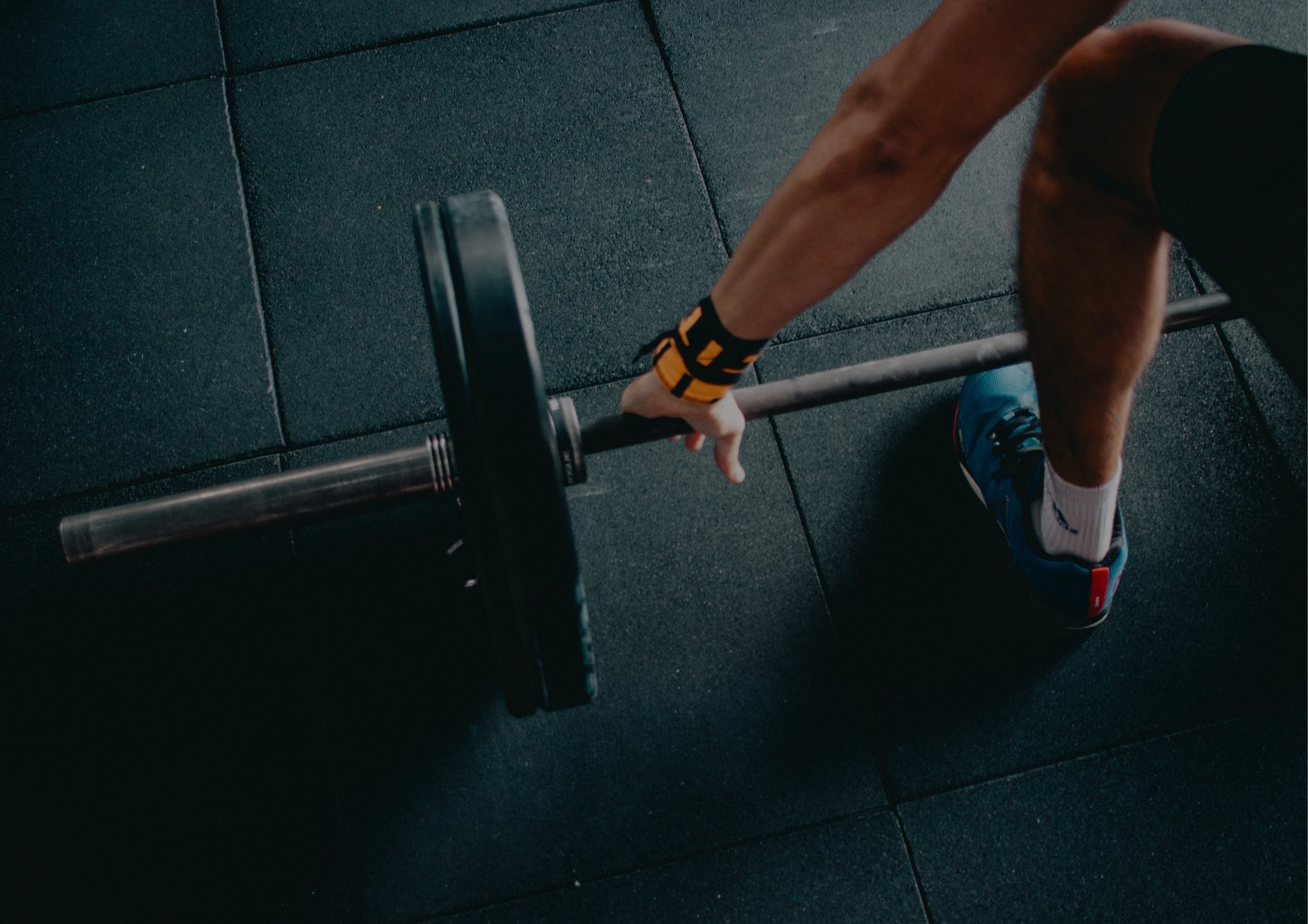 A person wearing athletic shoes and a wrist strap is reaching down to grip a barbell on a gym floor, preparing to lift the weight.