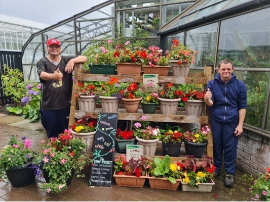 2 gardeners standing beside their re-stocked plant sale display featuring pots and hanging baskets.