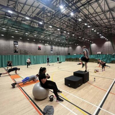 People taking part in a circuit‑style fitness class in a large sports hall, using equipment such as steps, mats, weights and an exercise ball across different workout stations.