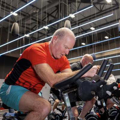 A group of people taking part in an indoor cycling class, riding stationary bikes in a large, modern fitness studio.
