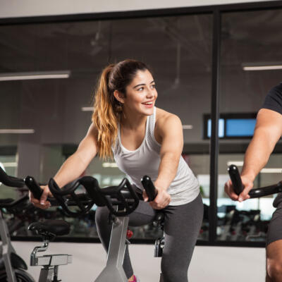 Two people riding stationary exercise bikes in a gym, leaning forward on the handlebars during a cycling workout.