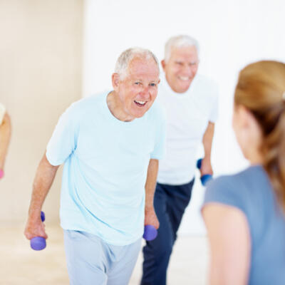 Smiling older adults holding small hand weights during an exercise class, with an instructor in the foreground.