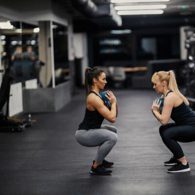 Two people performing weighted squats in a gym, each holding a small weight plate at their chest.