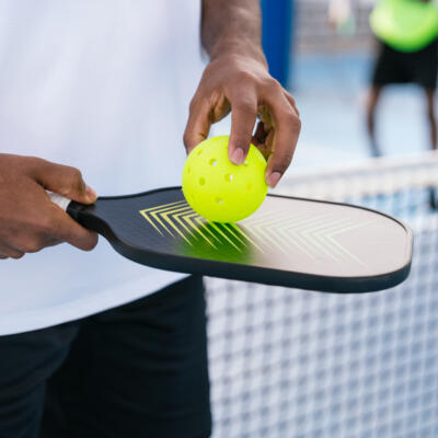 A person holds a yellow pickleball on a paddle at the edge of a court.