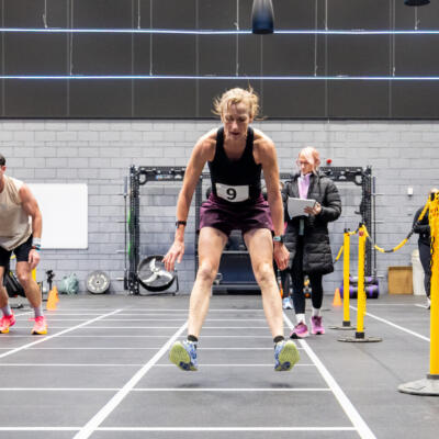 A person competing in an indoor fitness race, jumping forward between marked lanes while others and event officials watch in the background.