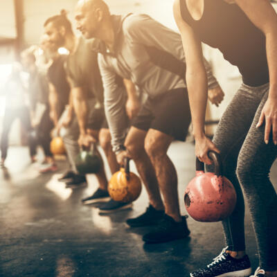 Group fitness class performing kettlebell exercises in a gym, with participants lined up and bending forward while holding colorful kettlebells on a dark floor.