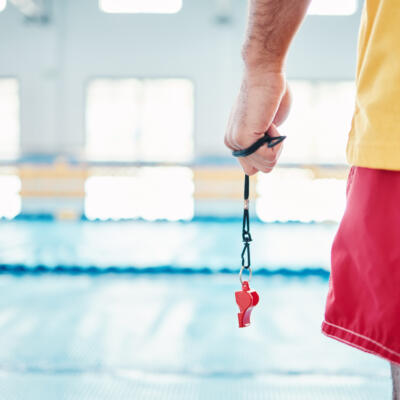 A lifeguard standing beside a swimming pool holding a whistle on a lanyard.