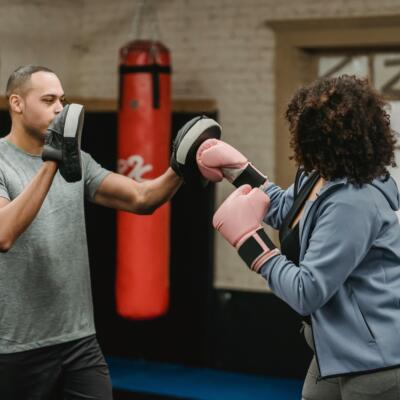 A boxing trainer holding focus pads while a person wearing pink gloves practises punches in a gym with heavy bags in the background.