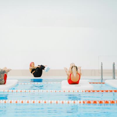 People taking part in a pool-based fitness class, balancing on floating mats while following an instructor at the pool edge.