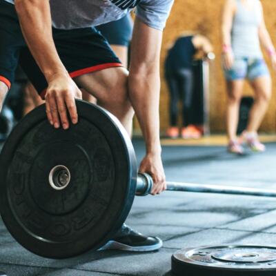 A person preparing to lift a barbell by adding weight plates in a gym, with others training in the background.