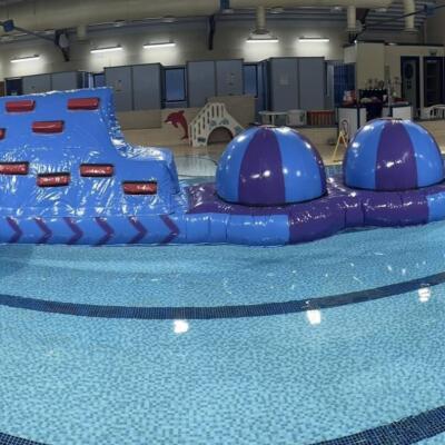 A large inflatable obstacle course floating in a swimming pool inside an indoor leisure centre.
