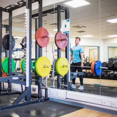 Person lifting a barbell in a gym with squat racks and wall mirrors.