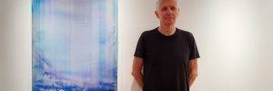 James Lumsden, artist, stands next to a painting hanging on the wall in an art gallery